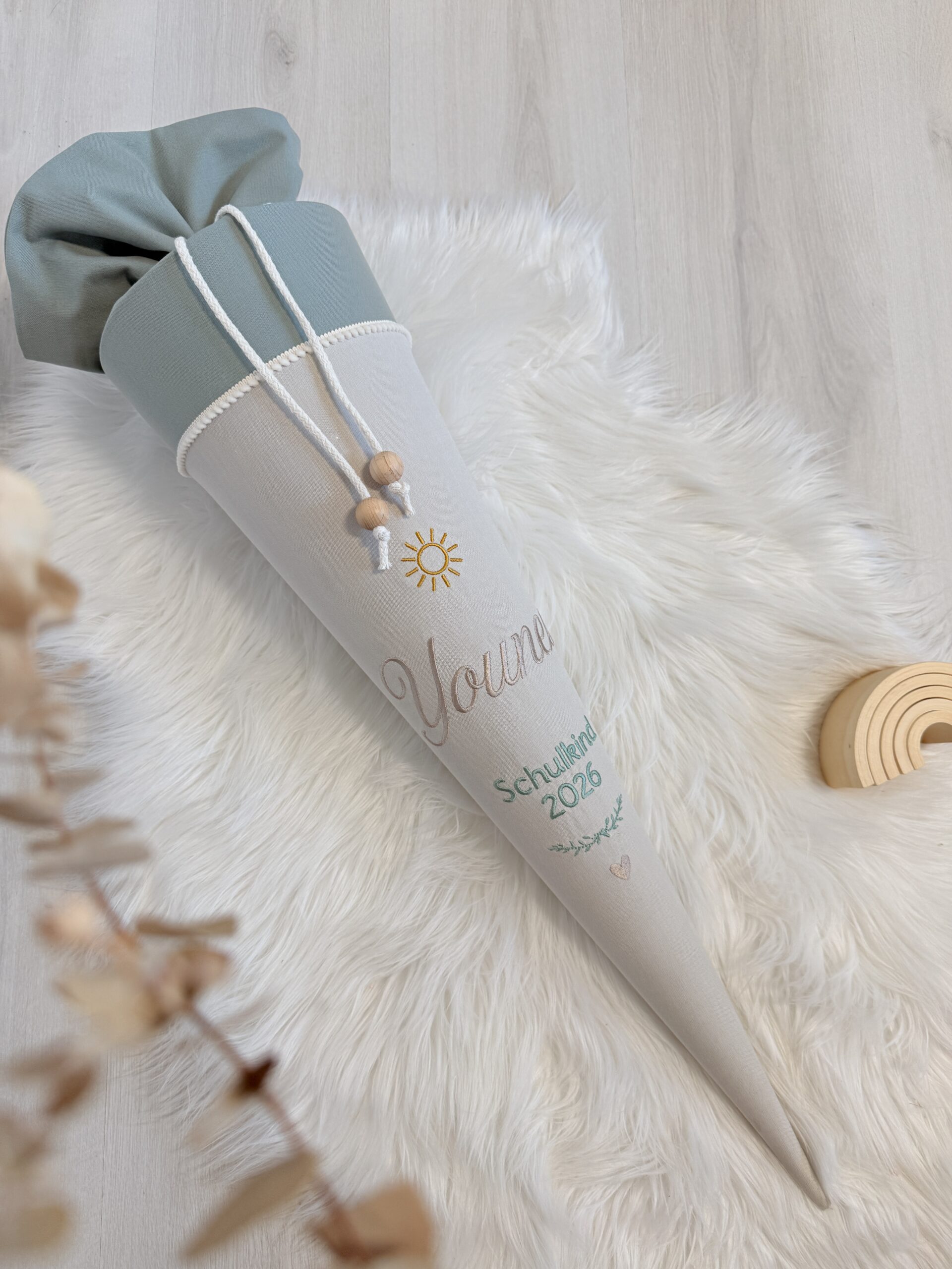 Beige-salbeifarbene Stoffschultüte mit Sonnenmotiv und Namensstickerei auf weißem Fell