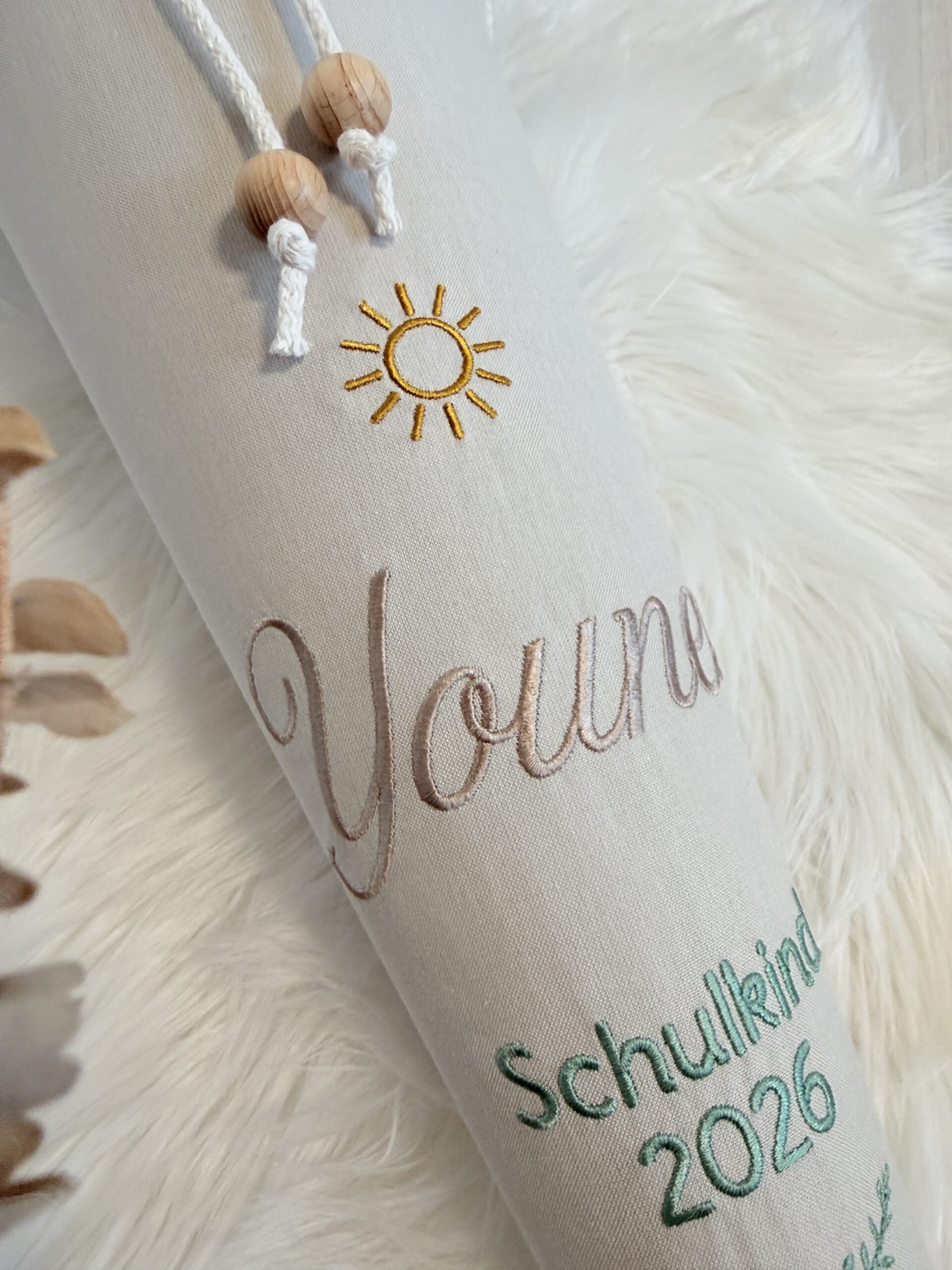 Nahaufnahme der gestickten Sonne und des Namens Younes unter weißen Kordeln mit Holzperlen